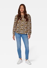 Eine Frau steht da und trägt eine blumige Langarmbluse, hellblaue Skinny Jeans und weiße Turnschuhe vor einem schlichten weißen Hintergrund.