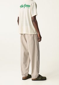 Osoba ubrana w luźne beżowe spodnie, kremową siateczkową koszulę z krótkim rękawem z zielonym napisem na plecach oraz zielone zamszowe klapki.