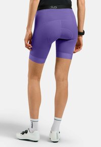 Lila Radsportshorts mit glatter Textur, hohem Bund und kontrastierenden Nähten. Kombiniert mit weißen Radsportschuhen und Socken.