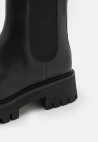 Botte en cuir noire avec semelle épaisse à crampons et panneau latéral élastique, montrée en gros plan sur fond blanc.