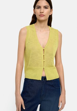 Kobieta w żółtej dzianinowej kamizelce bez rękawów z guzikami, dekoltem w serek i ciemnoniebieskich, wysokich dżinsach, stojąca na jednolitym tle.