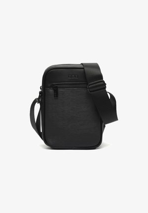 Malá černá crossbody taška s texturovaným předním dílem, zipovou kapsou a nastavitelným širokým popruhem na bílém pozadí.