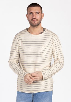 Mann mit kurzen Haaren, der ein beiges Langarmhemd mit dünnen grauen Streifen und blaue Jeans trägt, die Hände locker vor dem Körper verschränkt.