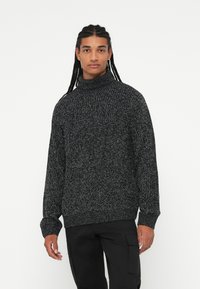 Chunky turtleneck striktrøje i mørkegrå med lysegrå prikker. Strikket tekstur, lange ærmer og ribbede kanter og manchetter.