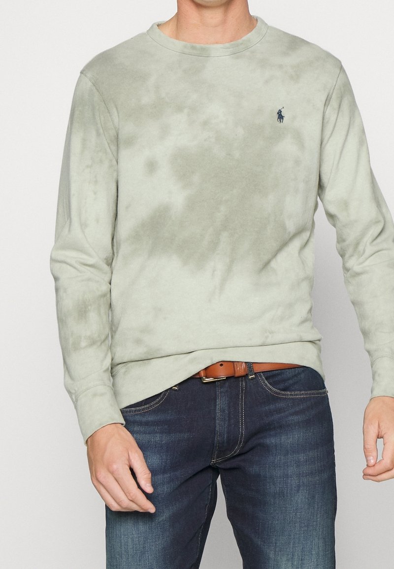 Homme portant une chemise tie-dye vert clair à manches longues avec un petit logo sombre, un jean bleu foncé et une ceinture marron, debout devant un fond uni.