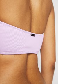 Top bandeau em lavanda com textura suave e uma pequena etiqueta preta na lateral. O design é minimalista e ajustado.