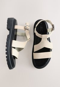 Beige lædersandaler med flere brede remme, justerbar spænde og sort gummisål med tekstureret slidmønster.