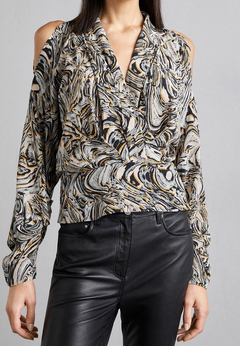 Femme portant un chemisier épaules dénudées avec un motif tourbillon noir, blanc et doré, associé à un pantalon en cuir noir taille haute.