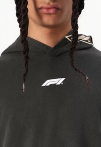 Čierna mikina s kapucňou a sťahovacou šnúrou, s bielym logom F1 na hrudi. Tkanina vyzerá hladko s jemným leskom. Viditeľné vrkoče.