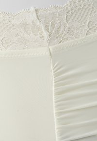 Lingerie marfim com um topo de renda texturizada e um tecido liso e pregueado abaixo. Costura detalhada nas bordas acrescenta interesse visual.