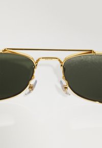 Ochelari de soare din metal auriu cu lentile verde închis, având un dublu pod și suporturi pentru nas. Accente clare pe vârfurile templelor adaugă detaliu.