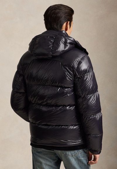Mann, der eine glänzende schwarze Steppjacke mit Kapuze trägt, von hinten gezeigt, kombiniert mit blauen Jeans vor einem schlichten beigen Hintergrund.