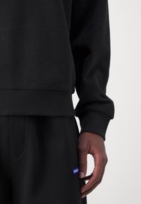 Gros plan d'une personne portant un pantalon de jogging noir et un sweat-shirt avec un logo HUGO bleu visible sur la poche du pantalon de jogging.