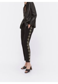 Vrouw in zwarte tapered broek met gouden en zilveren versieringen langs de zijkanten, gecombineerd met een zwart leren jack en zwarte haksandalen.