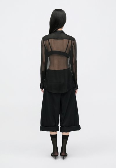 Black sheer long-sleeve shirt with a back panel design, paired with wide-leg black trousers, and black socks. Minimalist aesthetic.