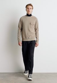 Felpa beige a girocollo con tasca sul petto, abbinata a pantaloni neri e sneakers bianche e nere. Il modello è in piedi su uno sfondo neutro.