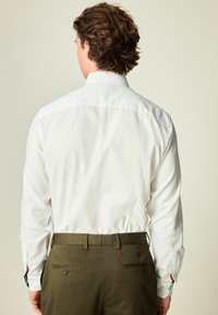 Camisa blanca de manga larga con textura suave, combinada con pantalones verdes oliva que tienen bolsillos traseros y puños interiores contrastantes.