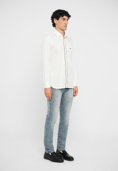 Vit knoppad skjorta med svart piping, lång ärm och en subtil logotyp. Pairad med ljusblå jeans och svarta oxfordskor.