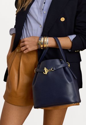 Námorná kožená taška typu vedro so zlatou kovovou prácou, so štruktúrovaným dnom, nastaviteľn�ým popruhom a textúrovaným povrchom. Skombinovaná s hnedou plisovanou sukňou.