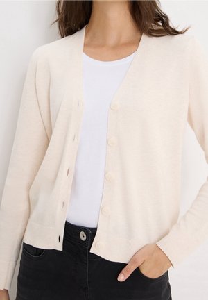 Femme portant un cardigan crème boutonné par-dessus une chemise blanche et un jean noir, avec une main dans la poche.