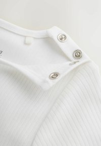 Tela blanca acanalada con tres botones metálicos de presión a lo largo de una costura del hombro, parcialmente abiertos para mostrar la textura interior y los detalles de las costuras.