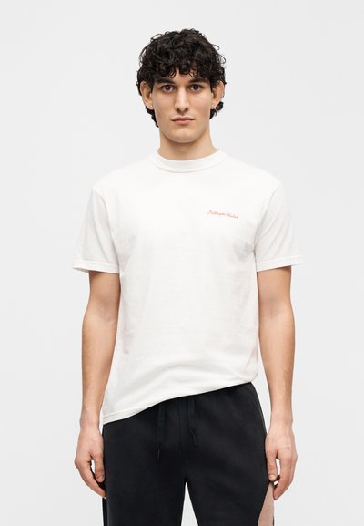 Λευκό βαμβακερό μπλουζάκι με στρογγυλή λαιμόκοψη και κοντά μανίκια· διαθέτει μικρό κόκκινο κεντητό κείμενο στην αριστερή πλευρά.