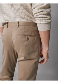 Pantaloni beige con una texture liscia, dotati di tasche posteriori e dettaglio a bottone, abbinati a un maglione crema con texture.