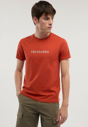 Giovane uomo che indossa una t-shirt arancione Trussardi e pantaloni cargo oliva, guarda di lato con le mani in tasca, sfondo neutro.