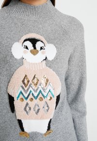 Grå stickad tröja med en pingvindesign i en rosa tröja, paljetterat geometriskt mönster och ribbat halsband.