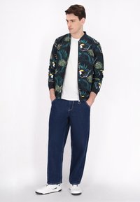 Bomberjack met een donker tropisch patroon, wit T-shirt, losse blauwe jeans en witte sneakers. Accenten zijn een rits en ribgebreide manchetten.