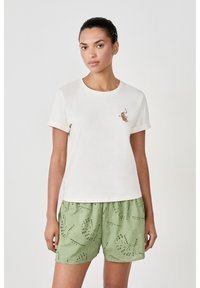 Camiseta de algodón de color beige claro con mangas cortas enrolladas y un pequeño diseño bordado en el pecho. Combinada con pantalones cortos verdes que presentan detalles de ojal.