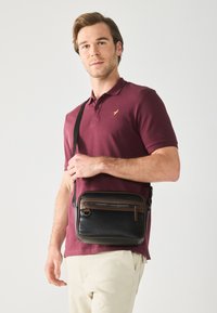 Sac messager en cuir noir avec une forme plate, des accents de zip marron et une bandoulière ajustable, porté à la main sous un polo bordeaux.
