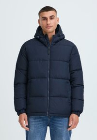 Marineblå puffjakke med aftagelig hætte, lynlåslukning, elastiske manchetter og quiltet design, båret over blå jeans.