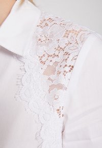 White blouse featuring lace detailing on the shoulder with floral patterns, textured fabric, and a classic collar.