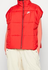Röd vadderad väst med hög krage, framzipper och Nike-logotyp. Ärmlös design, kviltad textur, parat med ljusa byxor.