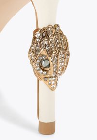 Hakken met een bovenwerk van witte leer en een gouden metalen decoratie met strass-steentjes en een centrale groene edelsteen op de hak.