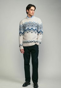 Pulover din lână crem cu modele geometrice albastre, cu mânici și tiv cu nervuri. Potrivit cu pantaloni închiși la culoare și pantofi, stând în fața unui fundal neutru.