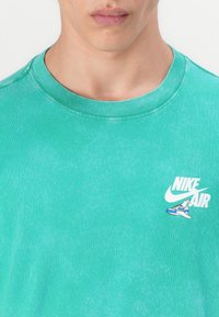 T-shirt de algodão verde menta com decote redondo, apresentando um pequeno logotipo e gráfico de sapato Nike Air em branco e azul no peito.