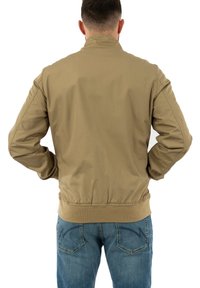Khaki bomber dzseki bordázott derékrésszel és mandzsettákkal, sima szövettextúrával. A hátsó nézet egyszerű, tiszta vonalakat mutat.