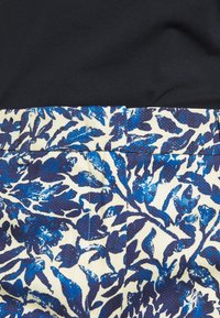 Blommiga byxor med en texturerad krämfärgad och blå design, med midjeband och sömdetaljer, delvis täckta av en svart topp.