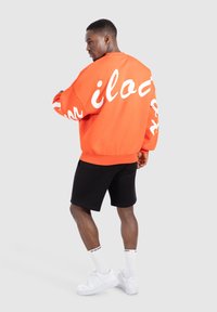 Felpa arancione con lettering bianco, vestibilità comoda, polsini a coste, abbinata a pantaloni corti neri, sneakers bianche e calzini bianchi.