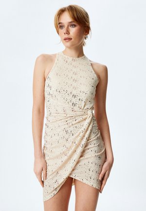 Robe à halter en beige clair avec un design froncé. Présente des speckles métalliques et une jupe en style enveloppant pour un look ajusté.