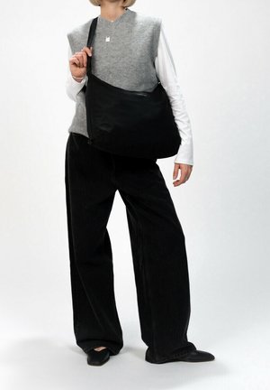 Černá nylonová taška přes rameno s nastavitelným popruhem, hladkou texturou, minimalistickým designem a zapínáním na zip, nesená osobou v neformálním oblečení.