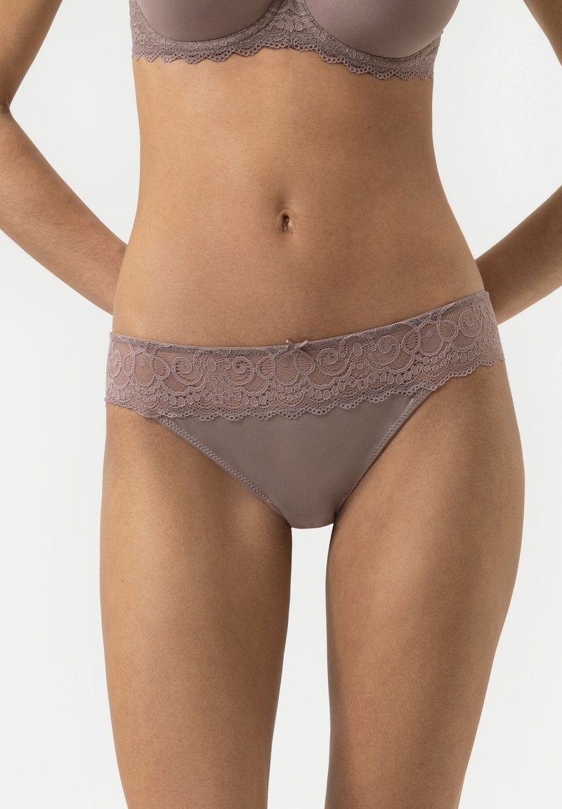 Woman wearing beige lace-trimmed bra and matching lace band bikini-style underwear, standing with hands behind back on white background.