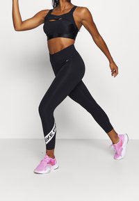 Czarny stanik sportowy z wycięciami, w połączeniu z czarnymi legginsami o wysokim stanie z białym logo "Nike Pro" oraz różowymi butami sportowymi.