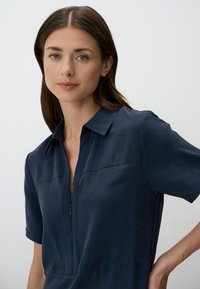 Robe courte bleu marine à manches courtes avec encolure à col et fermeture éclair à l'avant ; présente une texture lisse et un minimum de détails de couture.
