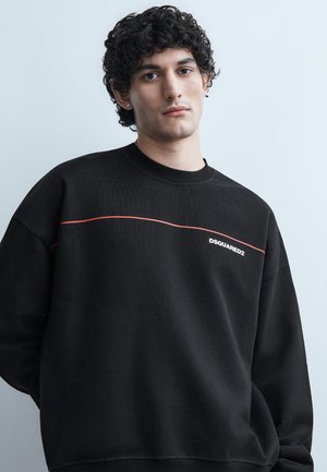 Νεαρός με κυματιστά μαλλιά που φοράει μαύρο φούτερ DSQUARED2 με λεπτή κόκκινη γραμμή στο στήθος, στέκεται μπροστά σε ανοιχτογκρι φόντο.