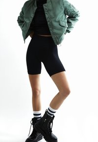 Groene bomberjack, zwart geribd cropped topje, zwarte biker shorts en zwarte combat boots met witte en marineblauwe gestreepte sokken. Minimalistisch ontwerp.