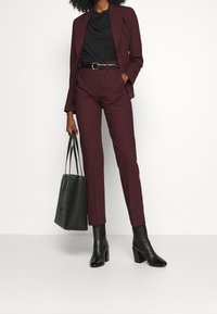 Bordowy garnitur z dopasowaną marynarką i spodniami z wysokim stanem. Czarna bluzka, pasek i botki za kostkę. Czarna torba na ramię z uchwytami. Gładka tekstura.