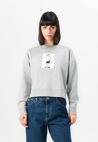 Donna con capelli scuri e lisci, che indossa un felpa grigia con una stampa fotografica in bianco e nero, abbinata a jeans blu larghi, su uno sfondo bianco.